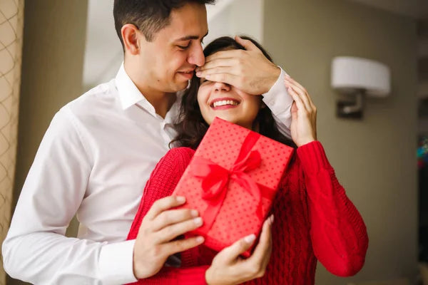 Man surprising a woman with a red gift box, both smiling indoors.